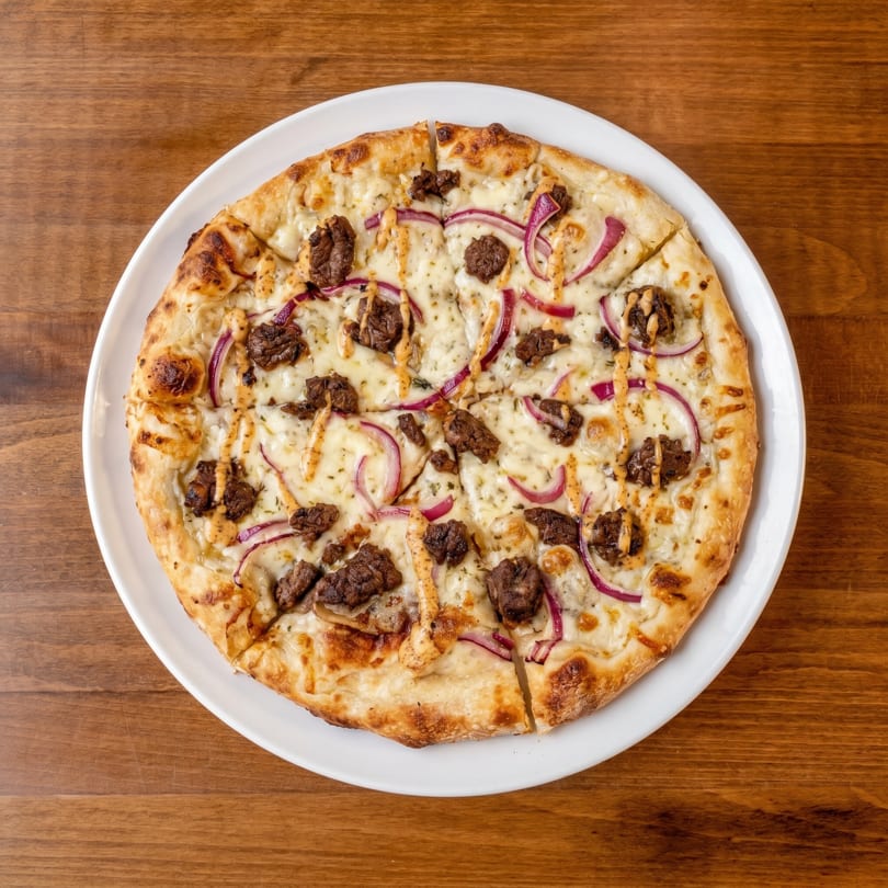Overhead view of a round pizza with white sauce, beef, red onions, and a drizzle of sauce.