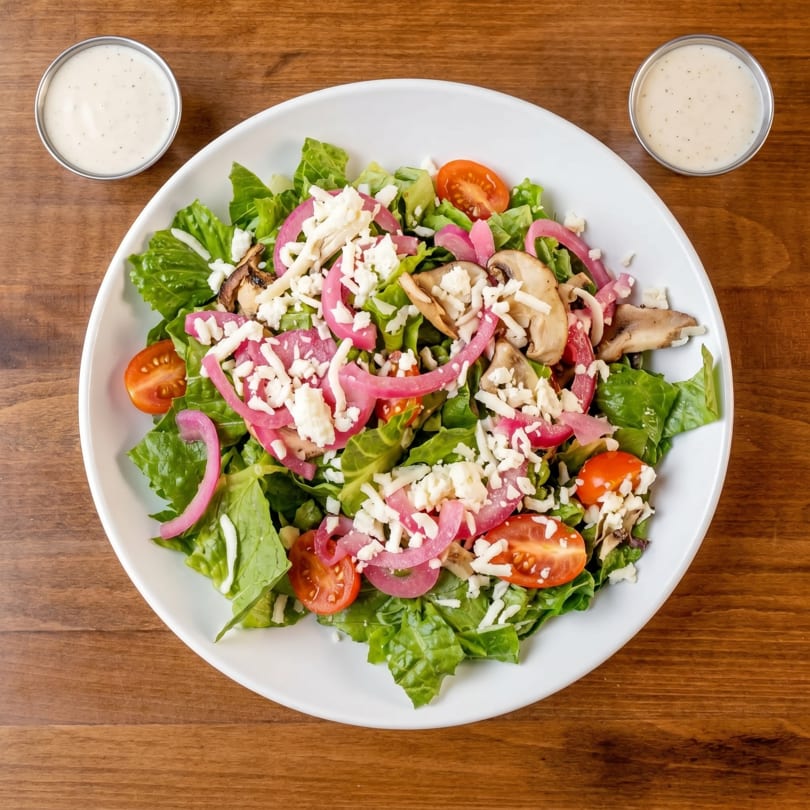 A fresh salad with lettuce, tomatoes, pickled onions, mushrooms, and crumbled cheese, with two dressing cups.