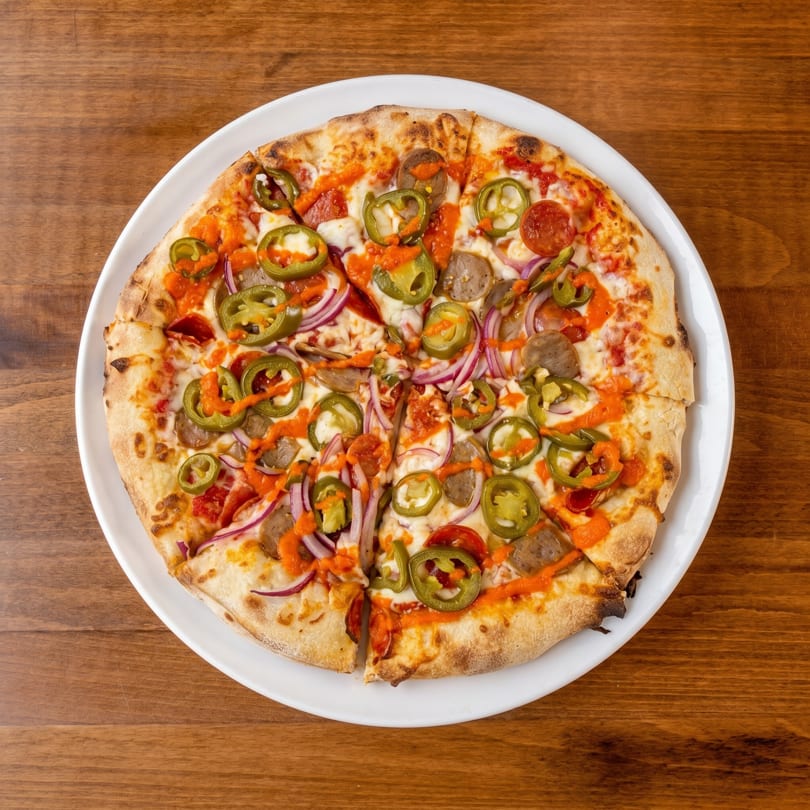 A sliced pizza with pepperoni, sausage, red onion, jalapeños, and orange sauce on a white plate.