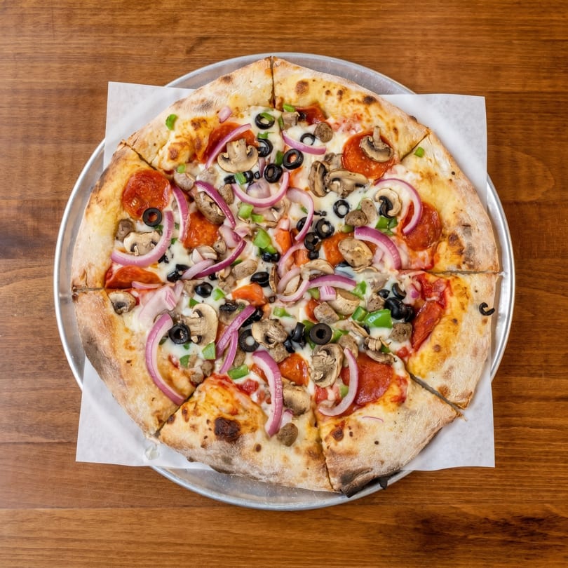 A whole pizza with pepperoni, mushrooms, olives, red onions, and green peppers on a wooden table.